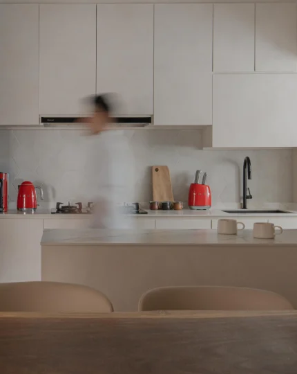 Compact white kitchen with counter and stools, black faucet, and a person in motion.